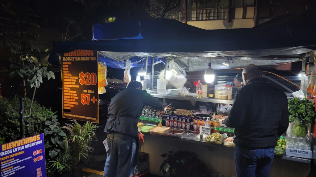 Puesto callejero con personas comiendo y una hilera de refrescos. Tacos estilo argentino uno de los lugares 24 horas en CDMX.