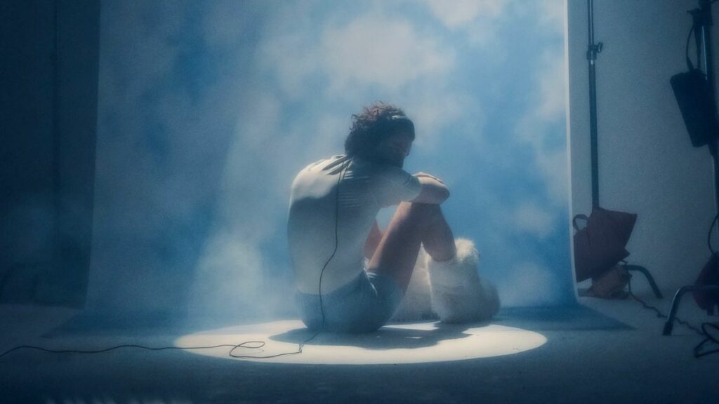 Humbe usando playera blanca, short azul y botas blancas con un fondo de nubes.