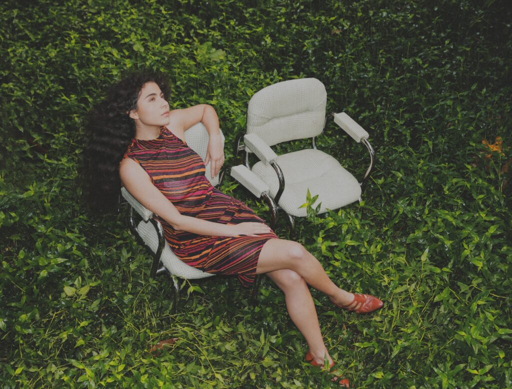 Silvana Estrada sentada en una silla en medio de un jardín usando un vestido naranja a rayas.