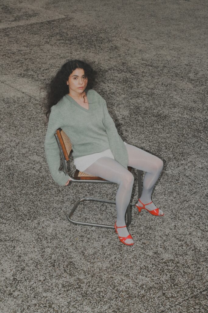 Silvana sentada en una silla usando un suéter verde, shorts blancos, medias plateadas y zapatos rojos.