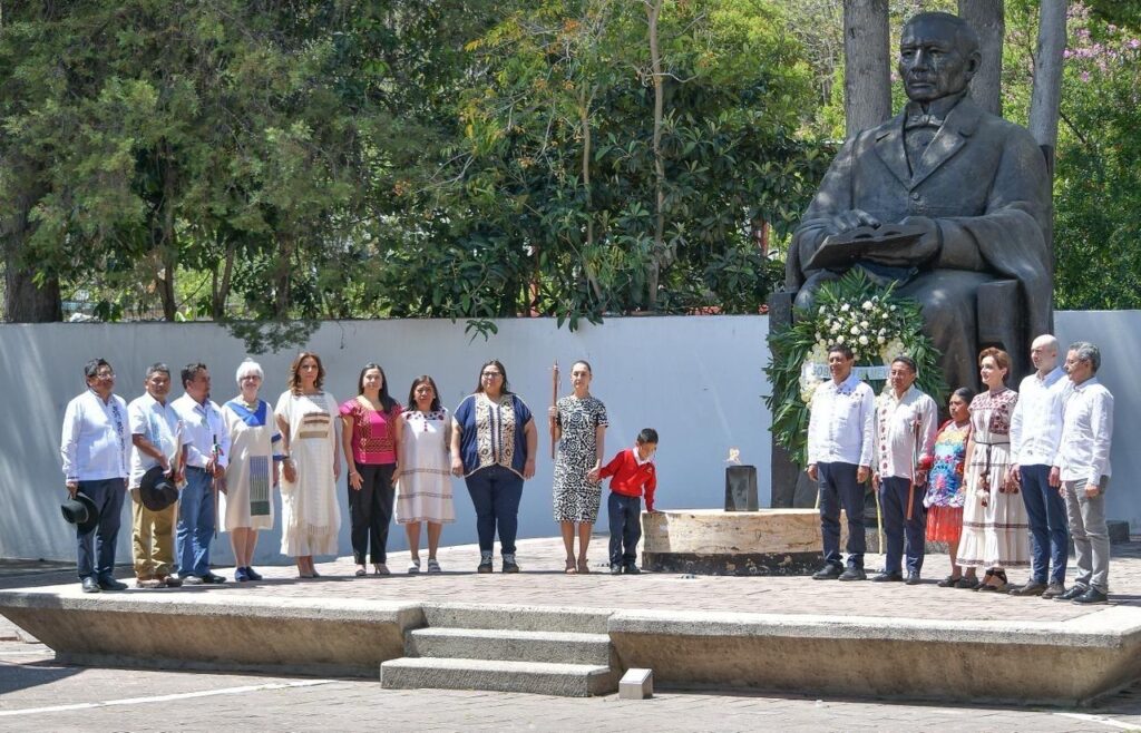 Fotografía de un grupo de personas de pie en un podio al aire libre, con una estatua grande detrás de ellos. La presidenta Claudia Sheinbaum está en el centro, y a su derecha, un niño con uniforme rojo. Un pequeño brasero con una llama está en el suelo.